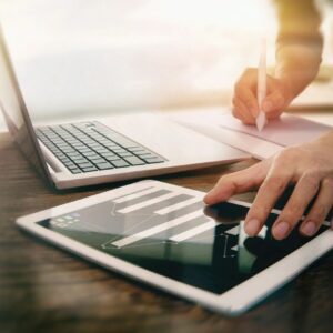Tablet and laptop with person writing notes.